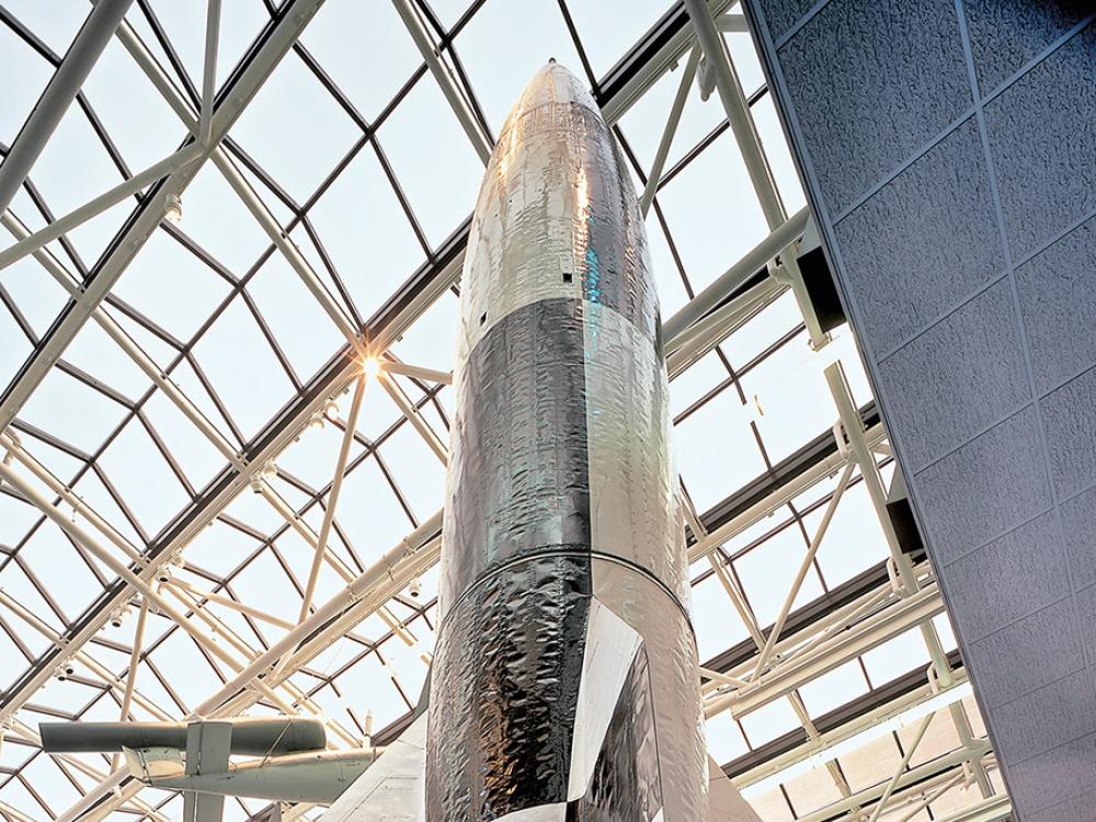 A V2 ballistic missile stands beneath a skylight in the Smithsonian.  Its cylindrical center section tapers through ogival sections to a warhead fuse and rocket engine exhaust in the tail, which has four swept fins with air vanes on the outer tips.