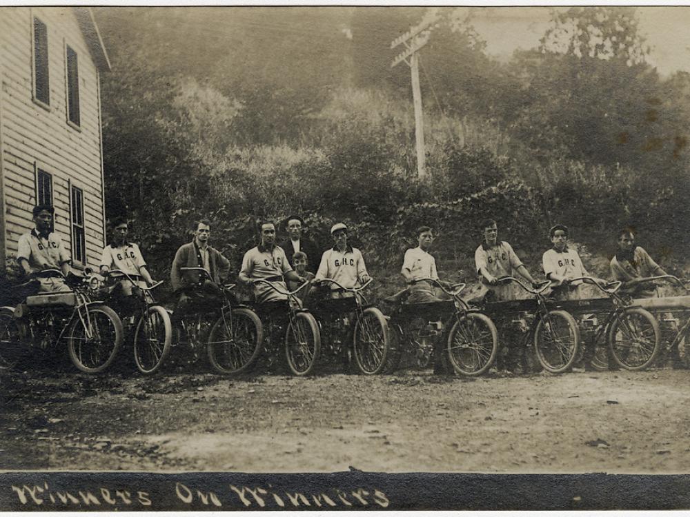 Eleven people pose with motorcycles in front of a building and bushes in this sepia-toned photo.