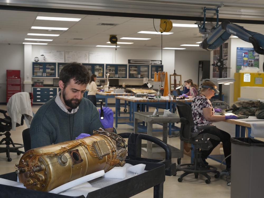 People at work in the conservation lab. In the foreground, a man holds a flashlight over a bronze cylinder with wires on it. In the background women sit at tables, the objects they're working on cannot be seen. 