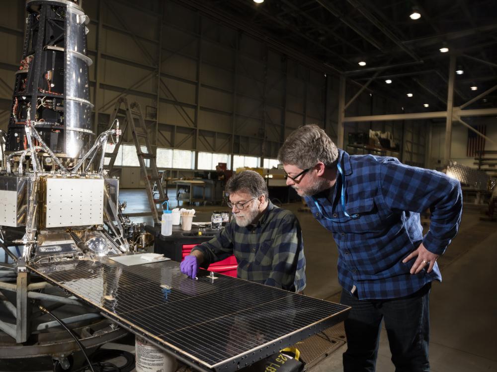 Two men lean over solar panel of lunar spacecraft undergoing restoration. 