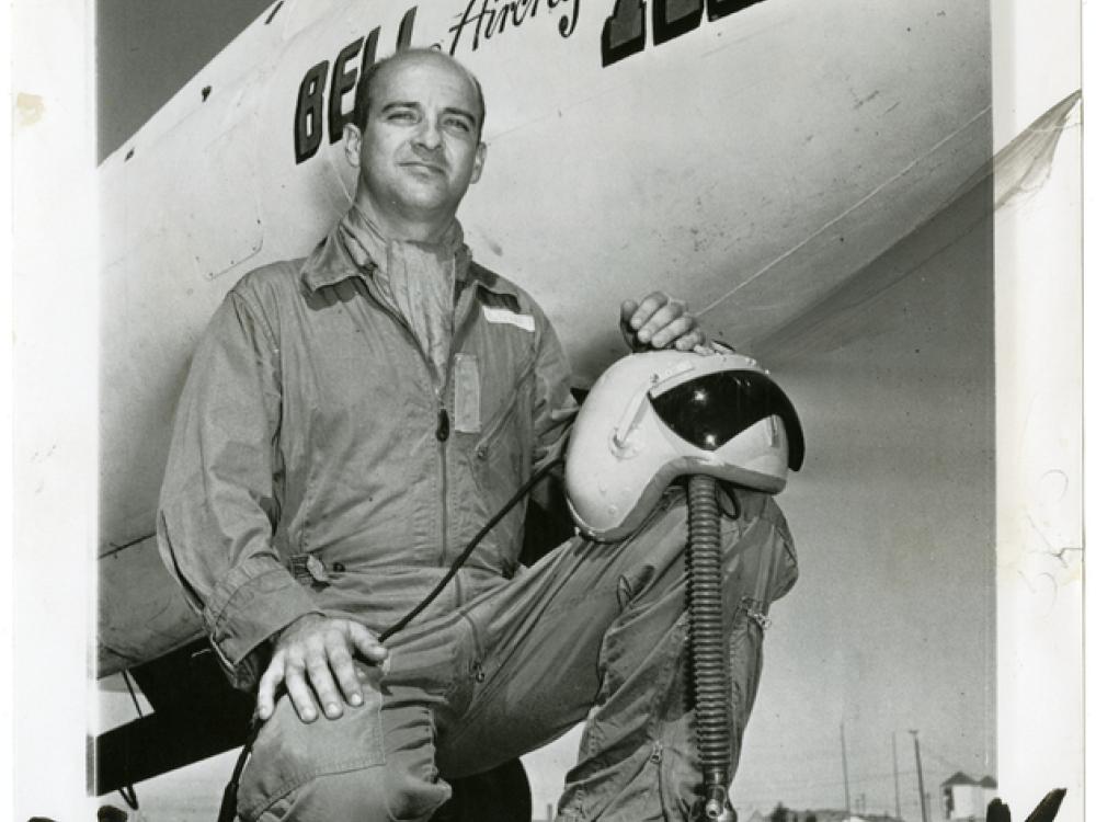 A man kneels in a flight suit in front of the nose of an aircraft which has Bell Aircraft X-2 written on the side. He balances a helmet on his knee.