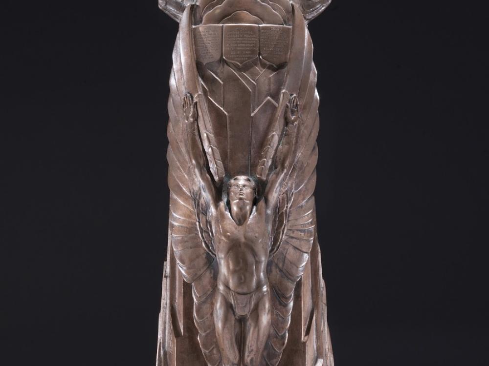 A bronze trophy with a male angel figure in front of a mountain with arms and wings stretched upward. Around the top half of the trophy are small plaques engraved with the details of each winner.