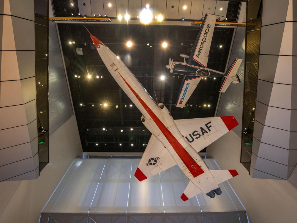 An orange and white plane with USAF written on the bottom of the wing seen from below. A smaller blue and white airplane hangs above it.