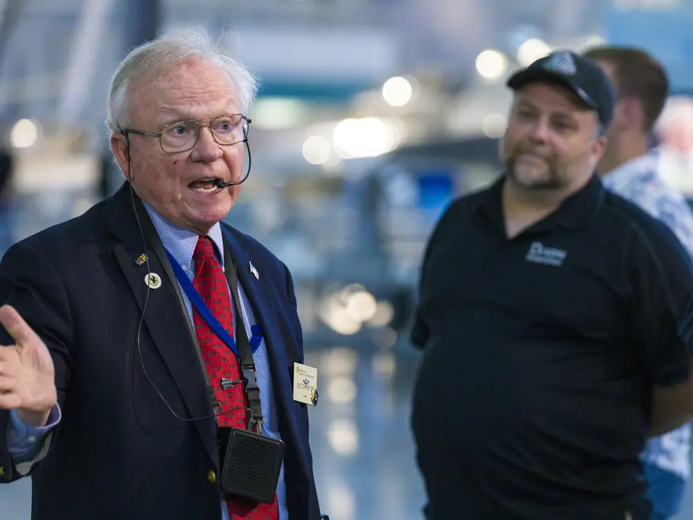 An older man with a speaker speaks and gestures while a middle aged man looks on. 