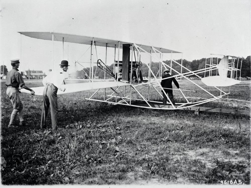 A photo of three men readying the Wright Military Flyer. Two men stand next to its wing, and the other stand between the landing skids.