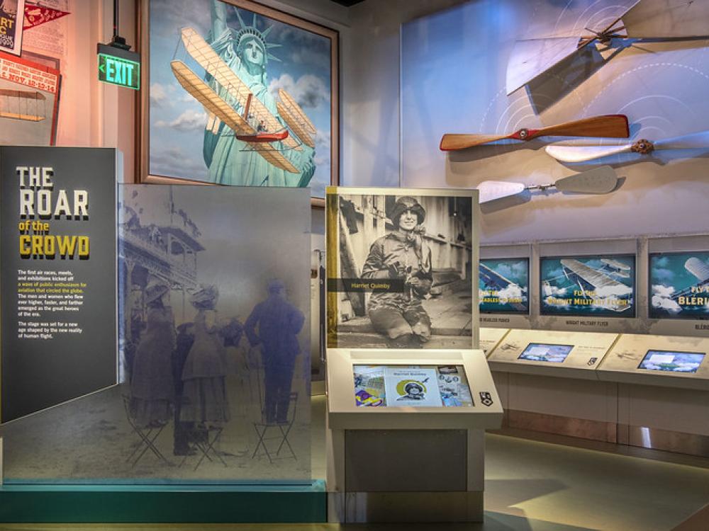 An image from inside the early flight exhibit. A panel says "The Roar of the Crowd" and there is a photo of Harriet Quimby. Propellers are hung on the wall.