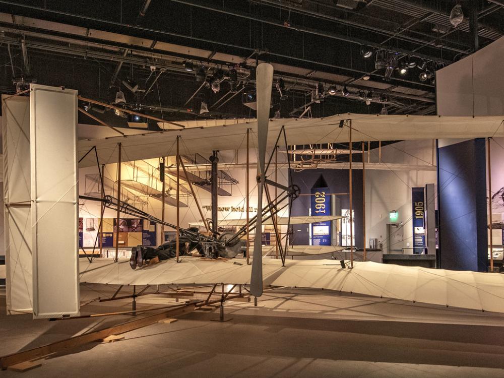 From behind, the view of the Wright Flyer in the Wright Brothers gallery. It is a beige biplane. 