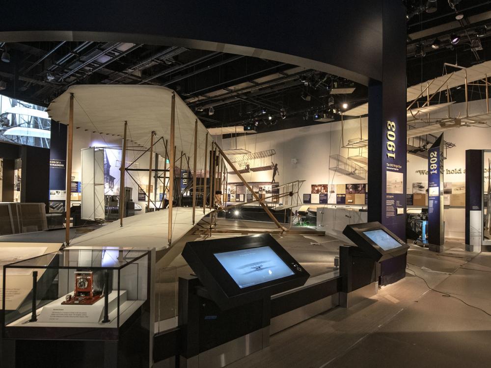 A view of the Wright Brothers gallery, with the Wright Flyer in the center surrounded by display panels and touch screens. 