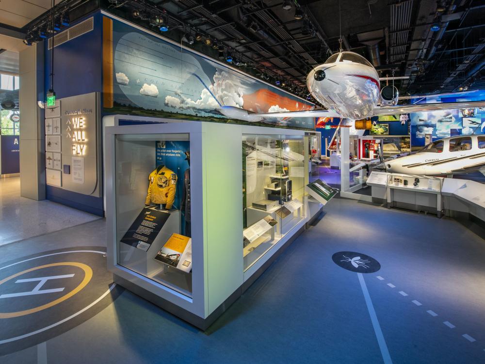A view of the We All Fly Gallery. A large airplane can be seen hanging overhead and a case with a jacket in it viewable from the ground. 