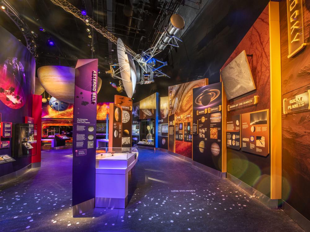 A view of the Kenneth C. Griffin Exploring the Planets Gallery. Th overwhelming colors are purple and orange. A model of the asteroid belt dots the floor. Pictures of space things line the walls. 