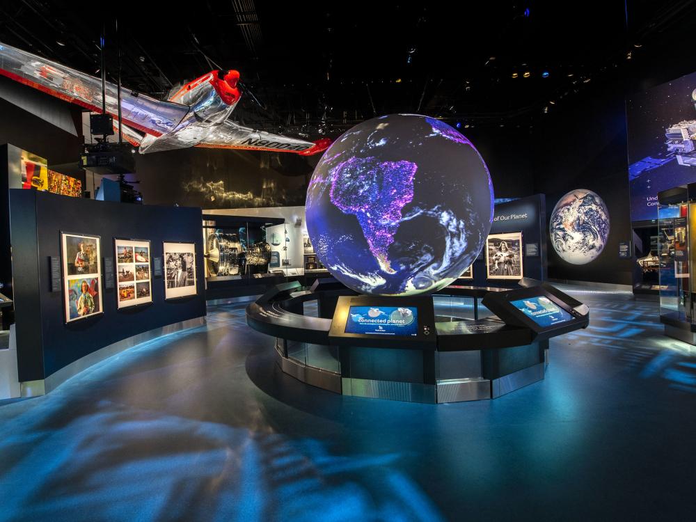A view of the One World Connected gallery. There is a large glowing globe visible at the center of the room, images around it, and a silver and red plane hanging overhead. 