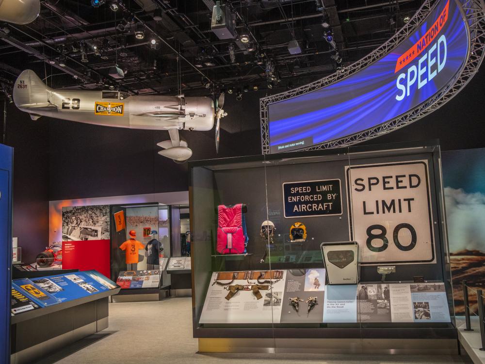 A photo of the inside of the Nation of Speed exhibit. A sign that says Nation of Speed hangs in the top right corner and across from it is Roscoe Turners airplane. A case with side signs that say speed limit 80 and speed limit enforced by aircraft is in the foreground.