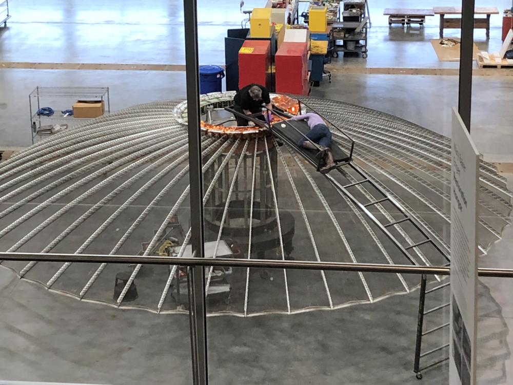 Two individuals work on the top of an umbrella shaped satellite in a restoration hangar.