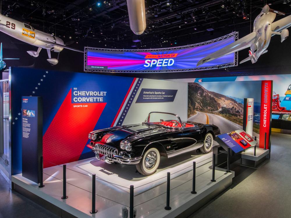 A photo taken from one of the corners of the Nation of Speed exhibit. Three aircraft hang from the ceiling in front of a sign that says Nation of Speed and a corvette is under the sign.