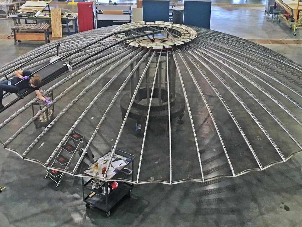 A person lying on top of a large umbrella shaped satellite antenna while cleaning it.