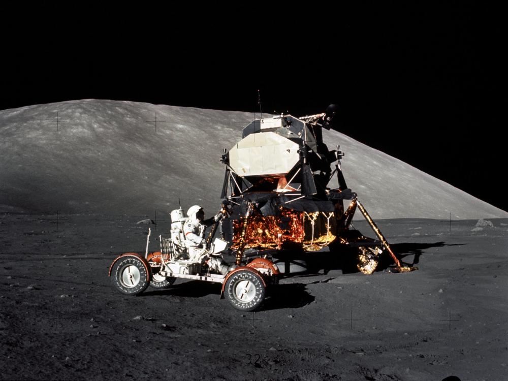 A lunar module and a lunar roving vehicle is photographed on the surface of the moon. An astronauts is sitting in the lunar roving vehicle.