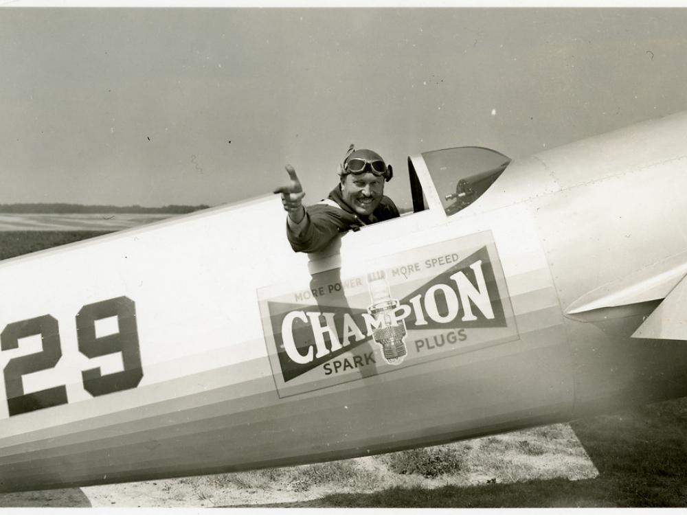 Side view of Roscoe Turner seated in the cockpit of his LTR-14 "Meteor" airplane on the ground. Turner points his right hand and winks.