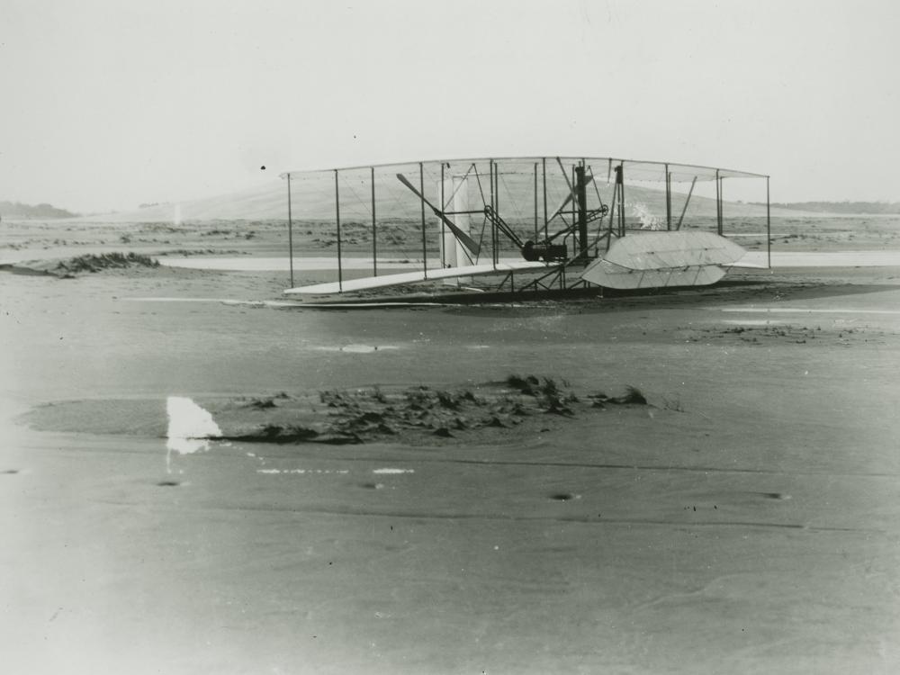 A black and white photo of a biplane on a sandy beach with a man standing the the far left. The front elevator is almost perpendicular to the plane as it is damaged.