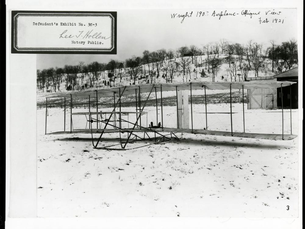 The Wright Flyer in the foreground of a snowy field, seen from the front. There is an exhibit number stamped at the top left.