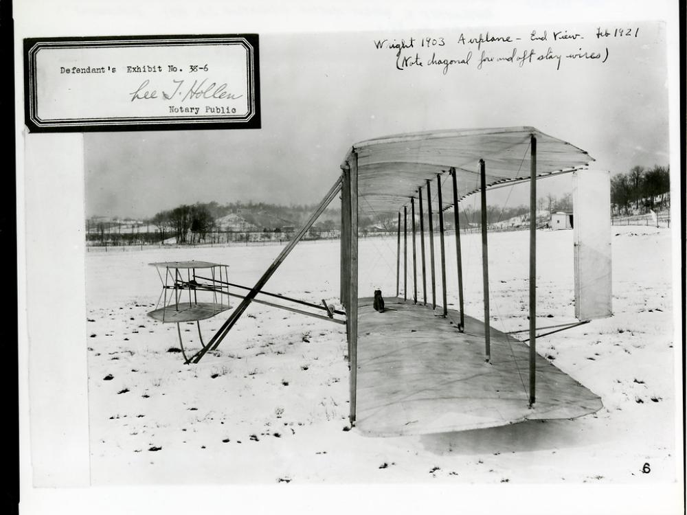 The Wright Flyer as seen from the side in a snowy field. There is an exhibit number stamped at the top left.