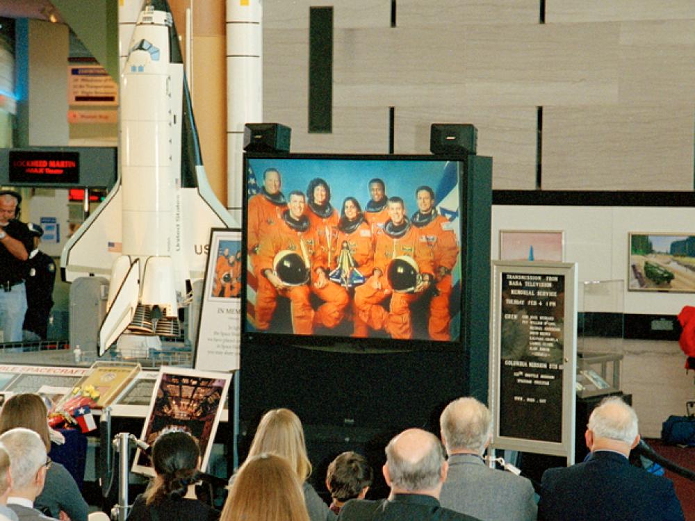 Astronauts are shown on a small tv screen while a small ground looks towards it.