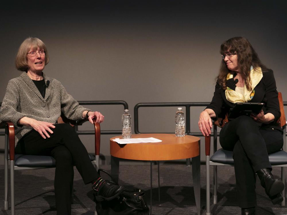 Two women sit in chairs. One speaks out to the crowd, the other is turned listening. 