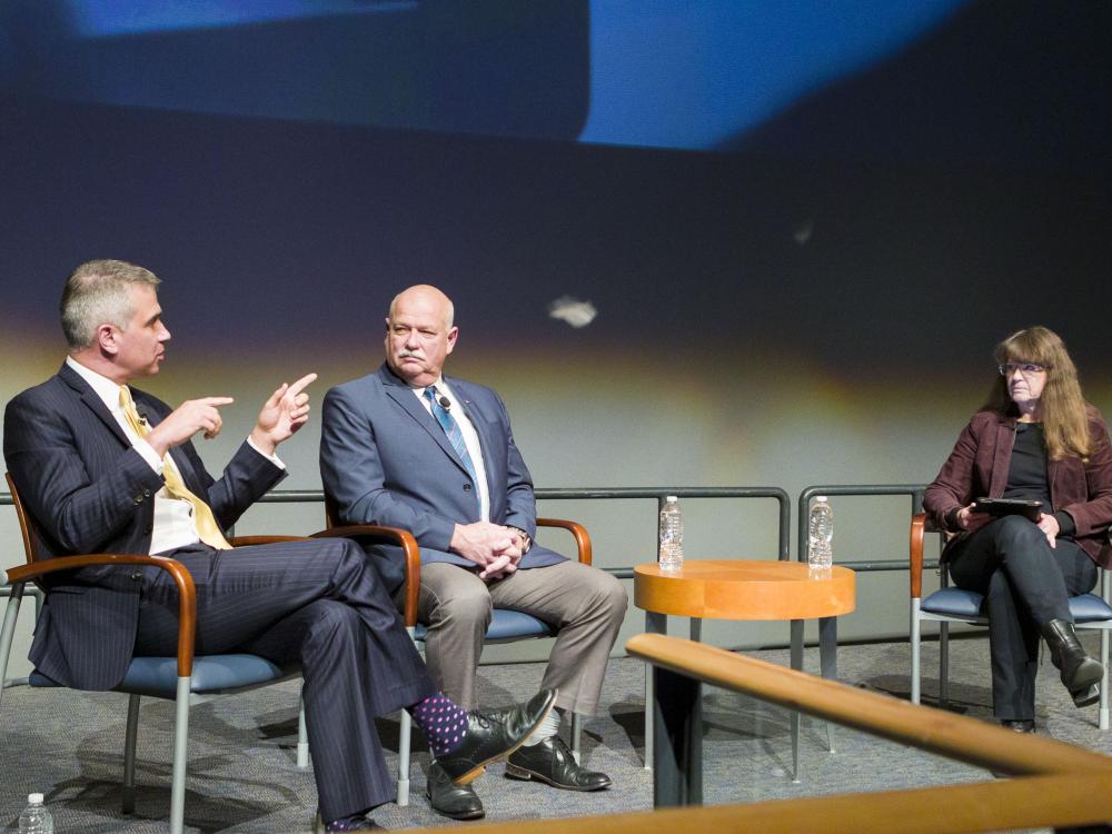 Three people sit on stage. One, a man, is talking and pointing. Another man and a woman sit looking at him. 