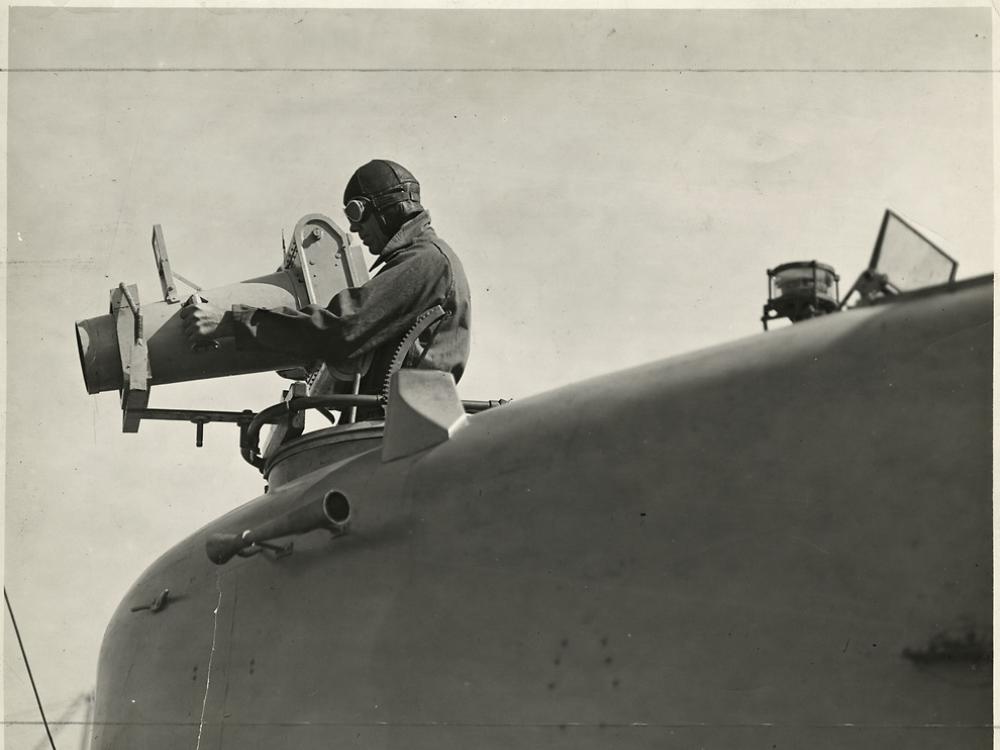 A close-up view from low left side of nose of aircraft, looking forward, of a man in a flight cap that stands in the forward cockpit of a Naval Aircraft Factory F-5L (PN-5) demonstrating a hand-operated K-2 aerial camera used for oblique aerial photography.