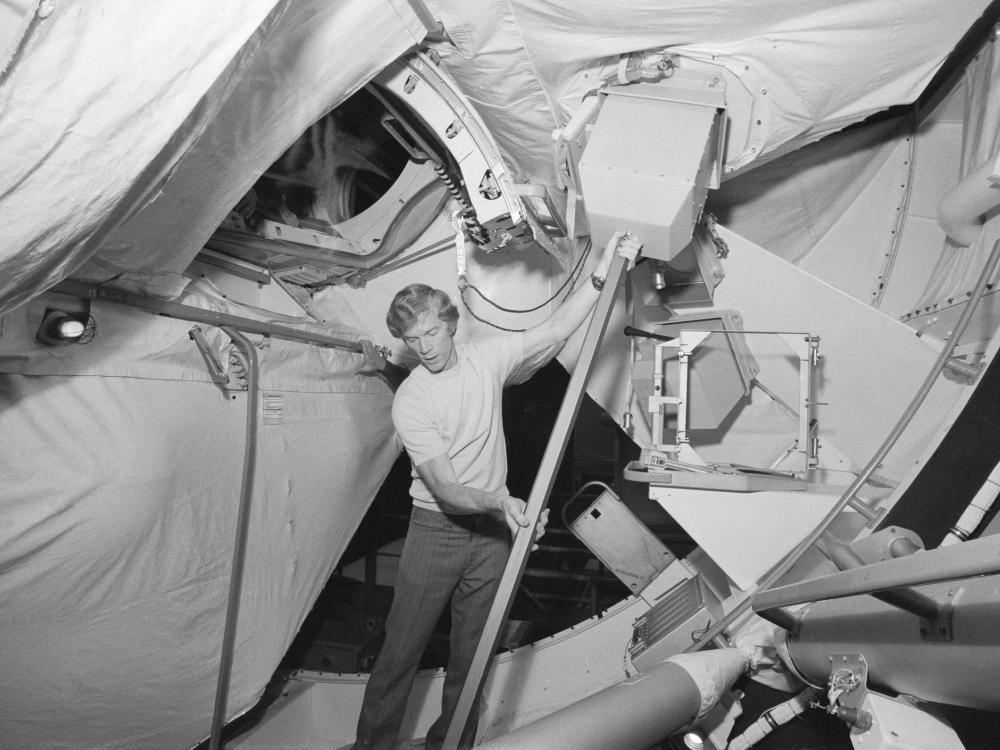 Black and white image of a man holding up a device in a space station simulator.