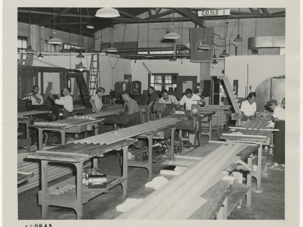 Black and white photograph of corrugated metal pieces with men and women working in the background