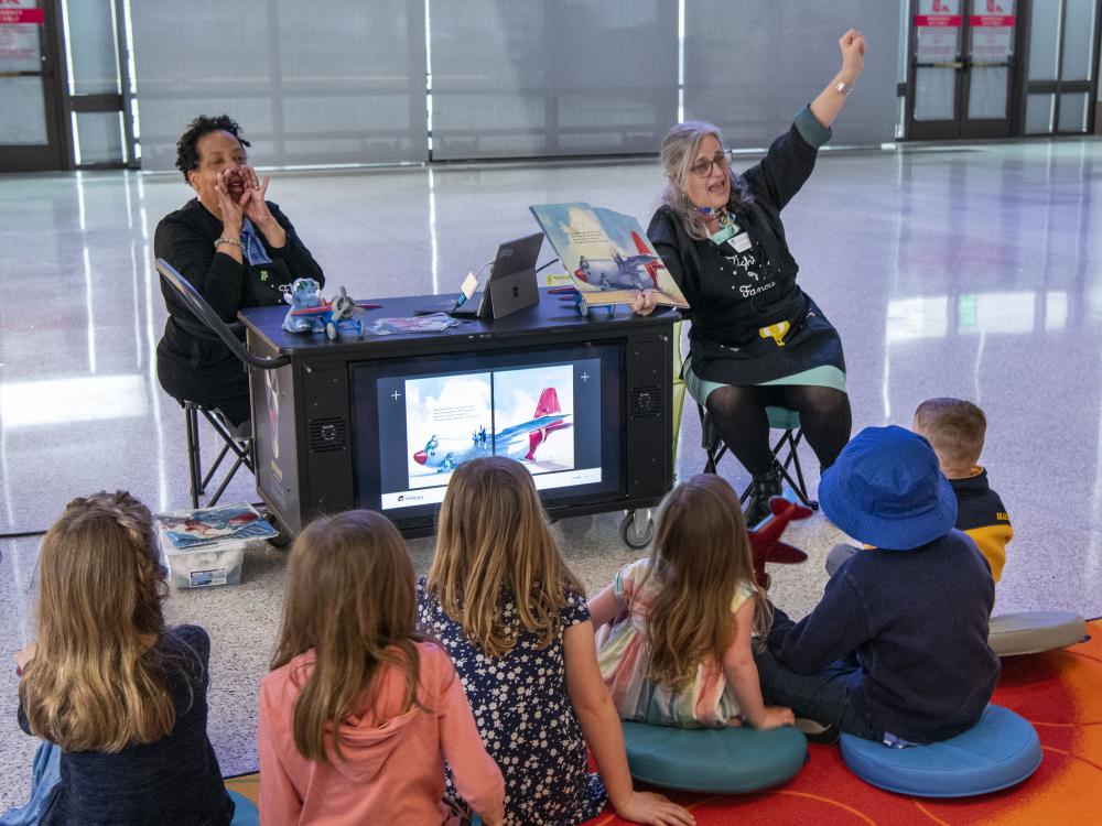 Two museum educators entertain a group of seated children. 