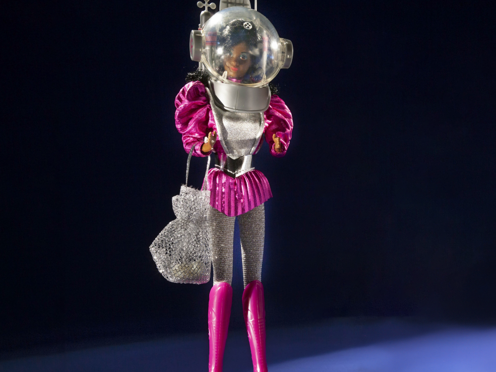 1985 Astronaut Barbie photographed against a dark background. She wears pink boots, silver leggings, a poofy pink top, and a space helmet.