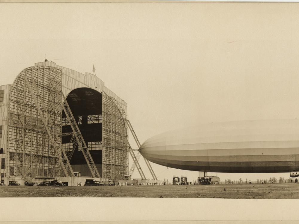  Panoramic photograph featuring a left side view of the US Navy airship Shenandoah under control of ground crew, being walked into its hangar.