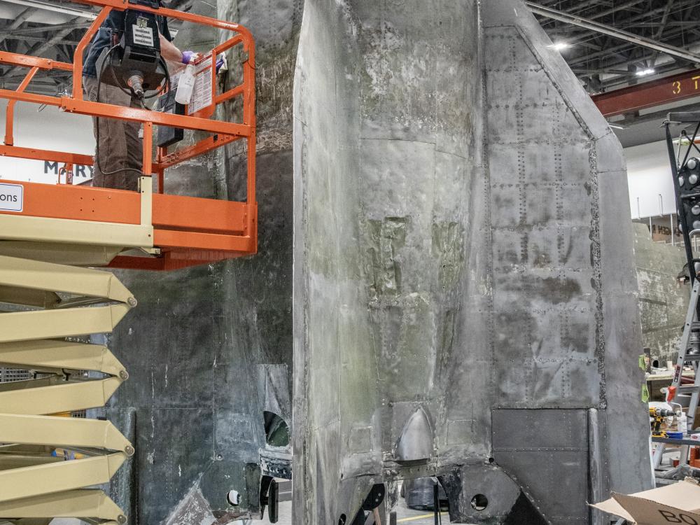 Large section of the bottom of a rocket is worked on inside a restoration facility.