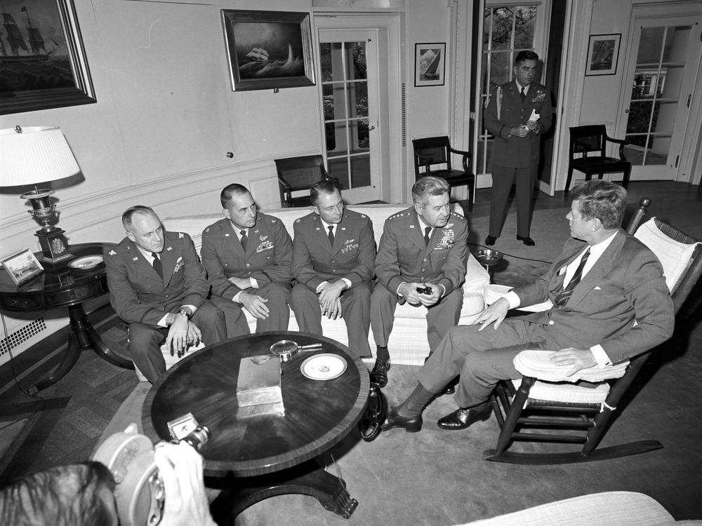 President Kennedy sits in a rocking chair, while four military pilots sit on a couch. 