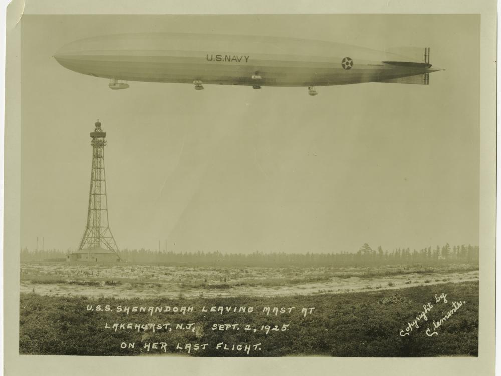 An airship floats in the air at the top of the frame. A mooring mast can be seen in the distance behind it.