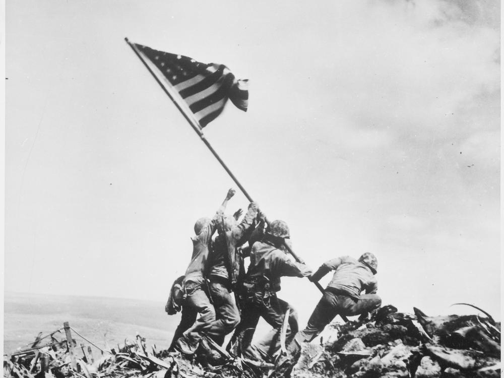 This iconic photograph shows four men in helmets raising an American flag over a pile of destruction. 