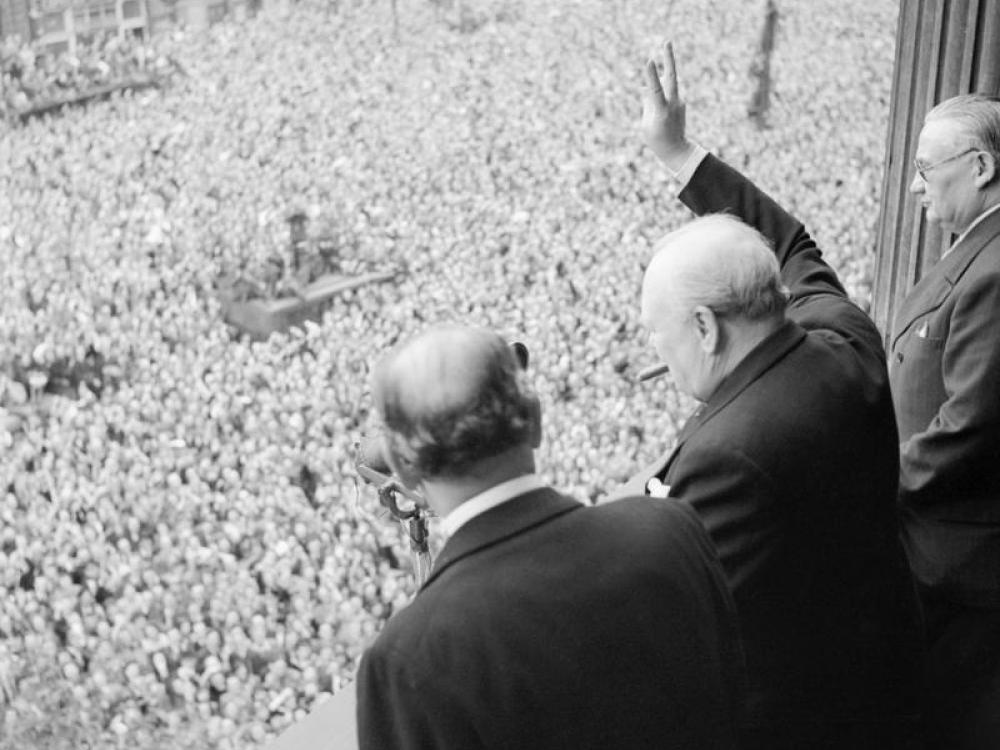 In this black and white photograph, three older men stand on a balcony overlooking streets full of hundreds of people. The one on the middle is clearly Winston Churchill--with his distinctive silhouette and cigar. He raises his had in a V for Victory gesture.
