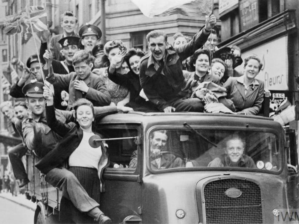 A truck overflowing with at least 20 people, some soldiers, some civilians, waving Union Jack flags, drives down the street. Everyone is jubilant. 