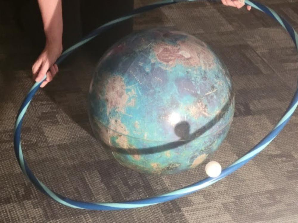 A hula hoop with a ping pong ball on it is held around a globe. 