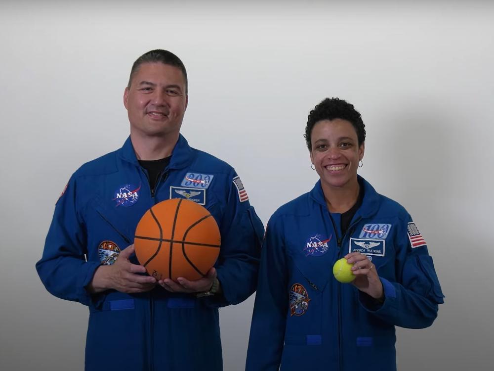 A tall man astronaut with a basketball and a shorter woman astronaut with a tennis ball. 