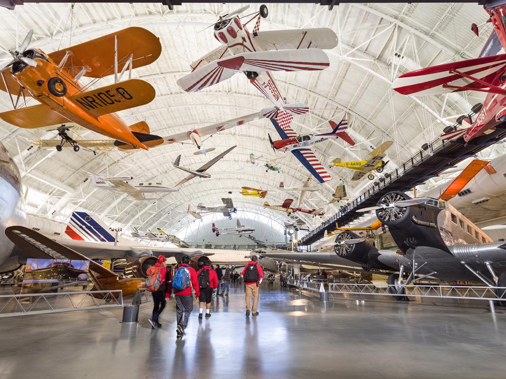 Planes both hand from the ceiling and stand on the ground in an aircraft hangar.