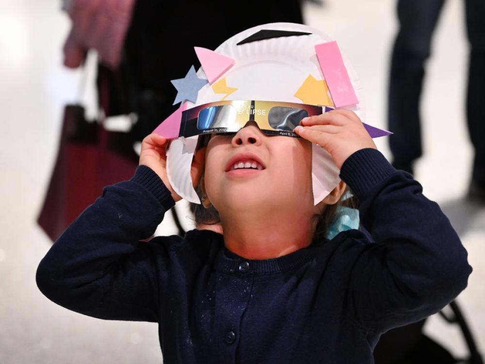A young visitor uses an eclipse viewer they made from eclipse glasses and a paper plate to safely view an annular eclipse. They squint up in concentration. 