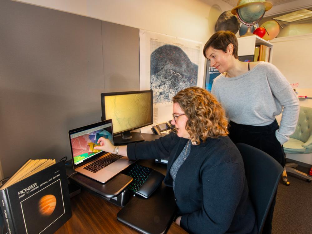 Two researches look at a computer monitor with colors displayed on it. In the background is a topographical map of a planet and globes of different planets. 
