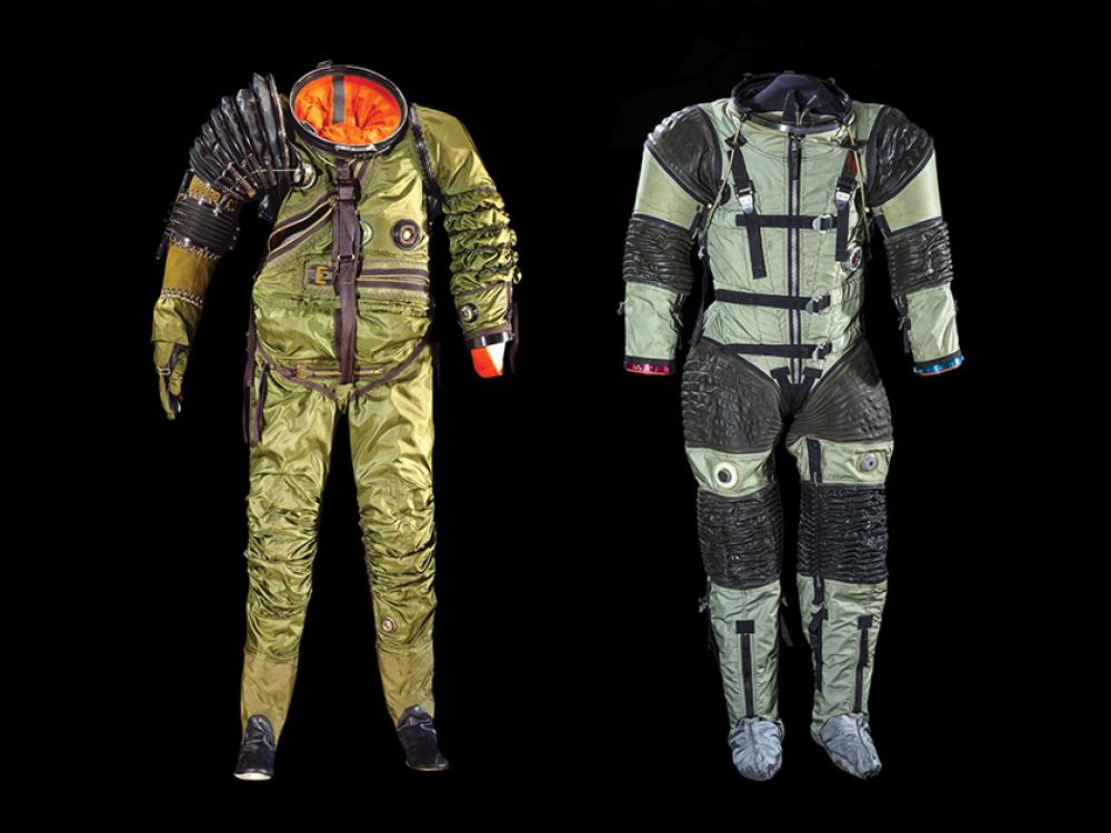 Left: An empty green spacesuit, with an orange interior peeking out through the top, has an oversized right shoulder that resembles a large accordian. Right: An empty light green spacesuit has black rubber material where a person's joints would be: shoulders, elbows, waist, and knees.