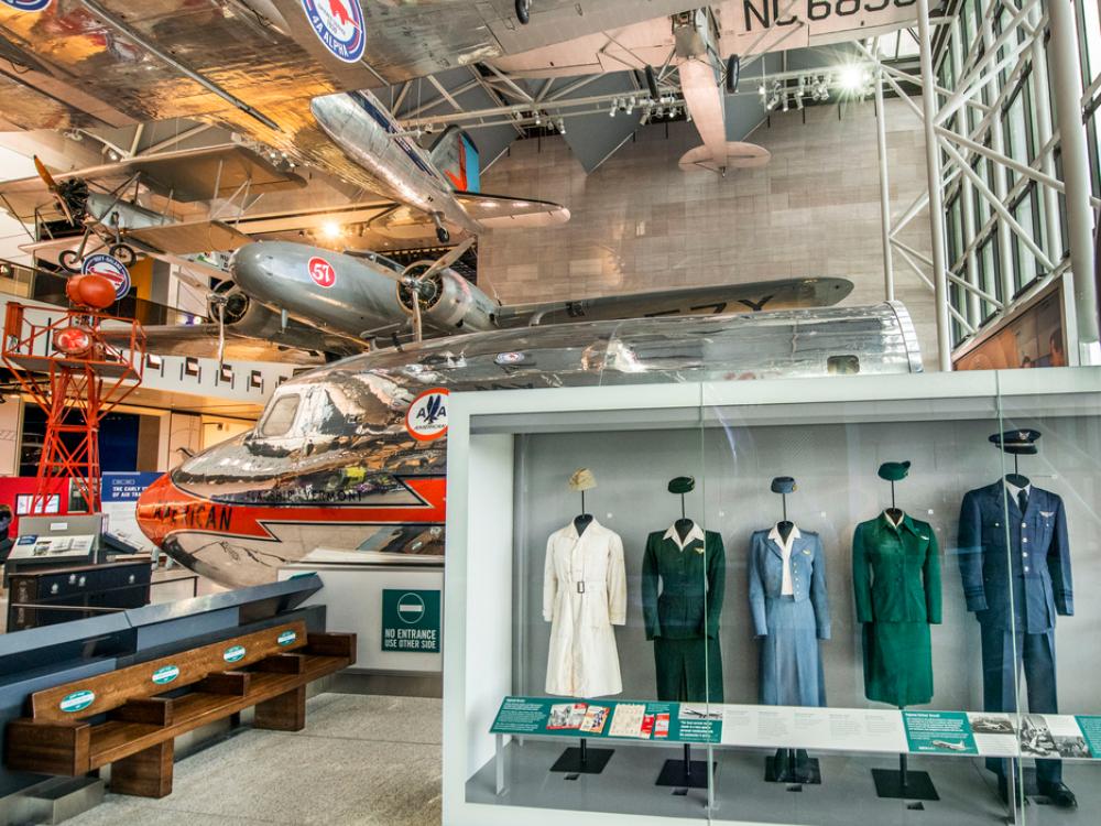 The exhibit America by Air as seen from the back wall. There are a row of flight attendant uniforms, a bench that shows the change is seat size over time, and the nose of a passenger airplane. 