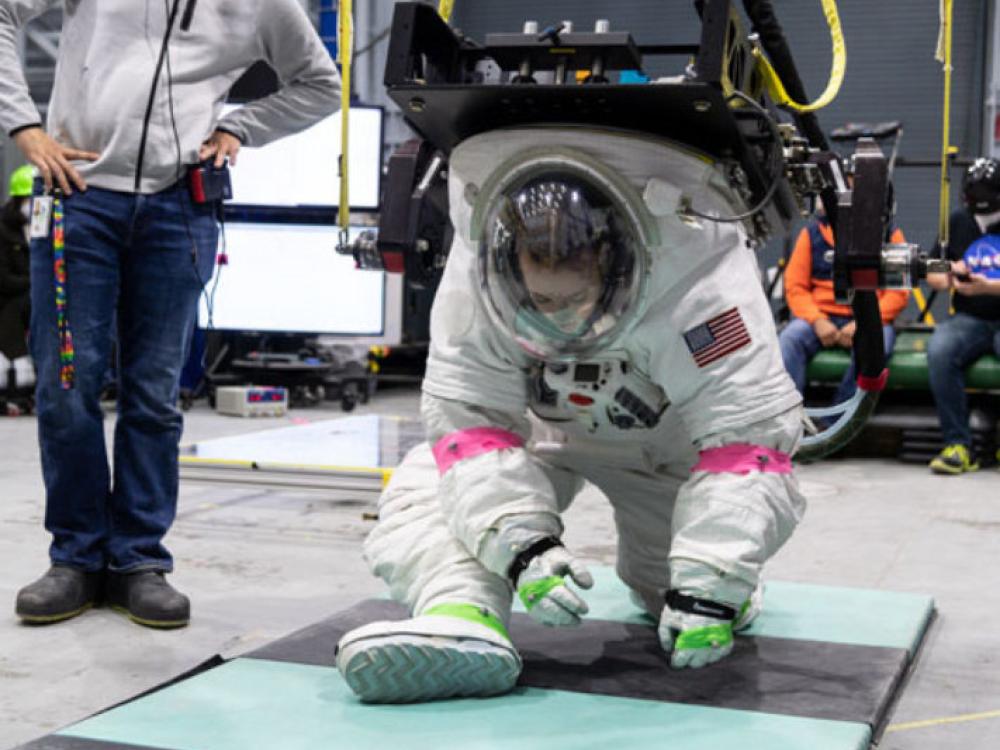 Christine Jerome wearing a NASA reference design suit for the newest spacesuits. She is crouched on the ground with one leg forward and one behind her. She appears to be pretending to pick something up from the ground. She is connected to an arm and some yellow tubes through a black rack on her back. A person is standing next to her, observing her movements.