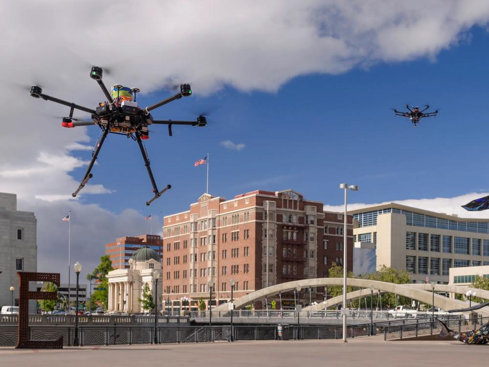Photo of two small drones flying over the capital city of Reno, Nevada, USA. Both drones look like they are either coming in for a landing or are just taking off. Each drone has 5 arms extending out from a central point, and each arm has an upward facing propeller at the end. One drone has two legs that look like landing gear, pointing down.