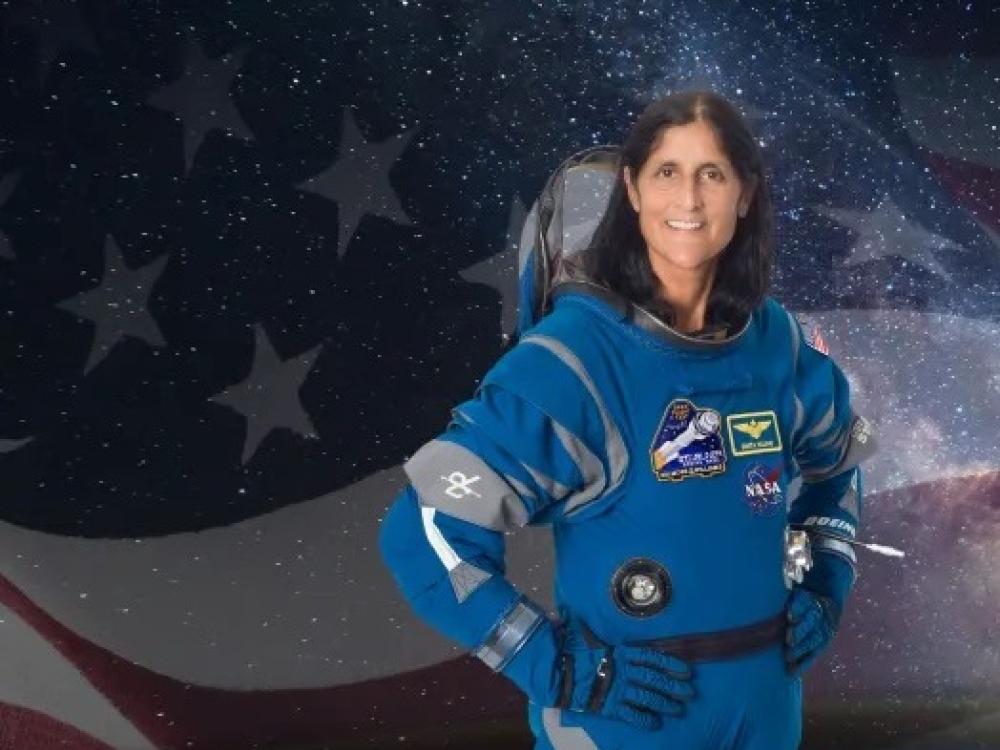 A woman in a blue spacesuit stands with her hands on her hip. The background is superimposed to be the American flag with the Earth underneath. 