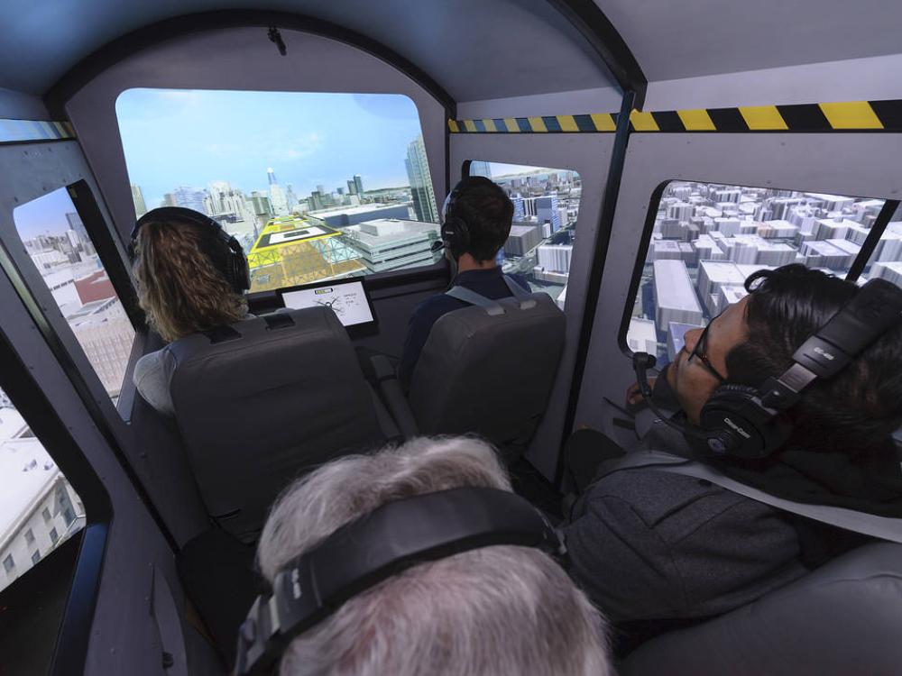 Photo of four people wearing headphones, riding in a simulator that looks like the inside of a small aircraft cockpit. The simulator has digital screens as windows and on the screens, it looks like the people are flying over a city. 